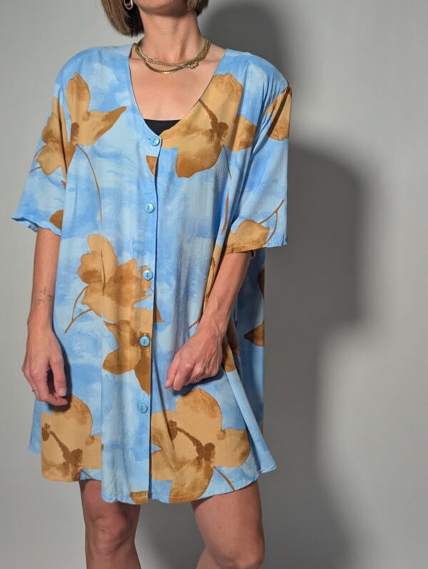 Vintage lichtblauwe blousejurk met grote bloemenprint in zand-, oker- en rozetinten, korte mouwen en knoopsluiting.