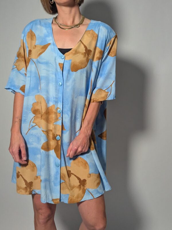 Vintage lichtblauwe blousejurk met grote bloemenprint in zand-, oker- en rozetinten, korte mouwen en knoopsluiting.