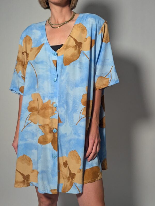 Vintage lichtblauwe blousejurk met grote bloemenprint in zand-, oker- en rozetinten, korte mouwen en knoopsluiting.