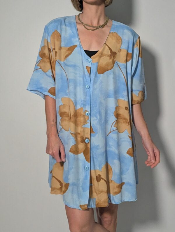 Vintage lichtblauwe blousejurk met grote bloemenprint in zand-, oker- en rozetinten, korte mouwen en knoopsluiting.